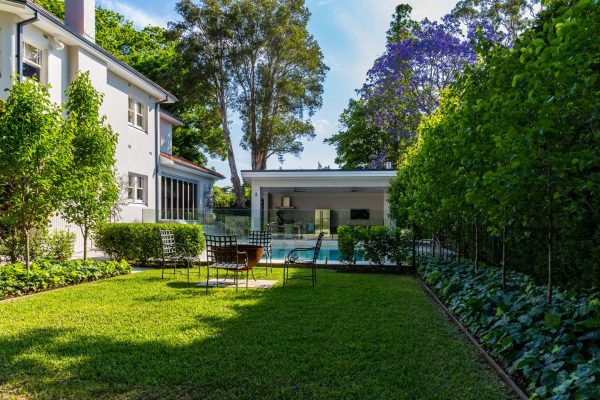 This example of Killara backyard landscaping includes an entertaining area on the lush grass complete with unique fire pit set. While the leaves and purple jacaranda flowers of long established trees dot the sky, the edges of the lawned area are lined with evenly spaced trees in formal orchard fashion, with full undergrowth.
